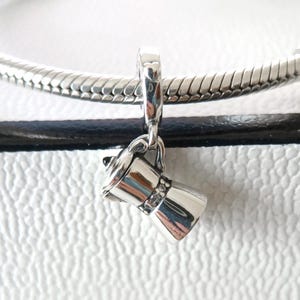 May include: A silver charm in the shape of a coffee maker, suspended from a silver chain. The charm has a detailed design, including a handle and a spout. The charm is likely made of sterling silver.