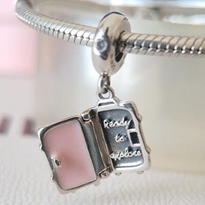 Puede incluir: Un charm de plata con un diseño de maleta. La maleta está abierta, revelando un interior rosa. El texto "Ready to Explore" está grabado en la maleta. El charm está unido a una pulsera de plata.