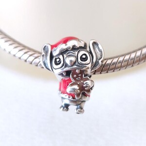May include: A silver charm featuring a cartoon character wearing a red Santa hat and jacket, holding a gingerbread man. The charm has black eyes and is on a silver bracelet.