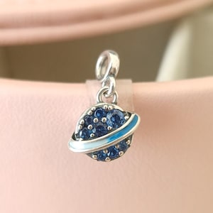 May include: A silver charm featuring a planet design. The planet is adorned with dark blue gemstones and a light blue and white band. The charm has a loop for attaching to a bracelet or necklace.