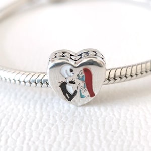 Puede incluir: Un charm de plata en forma de corazón, con una imagen pintada de Jack Skellington y Sally de Pesadilla antes de Navidad. El charm está unido a una pulsera de plata, con un diseño detallado en el borde del corazón.