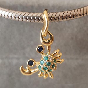 May include: A gold-toned crab charm hanging from a silver-toned snake chain bracelet. The crab features two dark blue circular eyes and a shell encrusted with small, sparkling teal and clear stones, finished with polished gold-toned claws and legs.