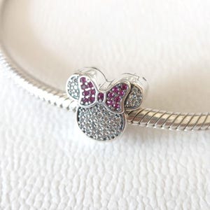 Op de afbeelding: Een zilveren bedel in de vorm van een muizenkop met een strik. De strik is rood en de kop is bedekt met kleine, heldere en rode steentjes. De bedel zit aan een zilveren armband.