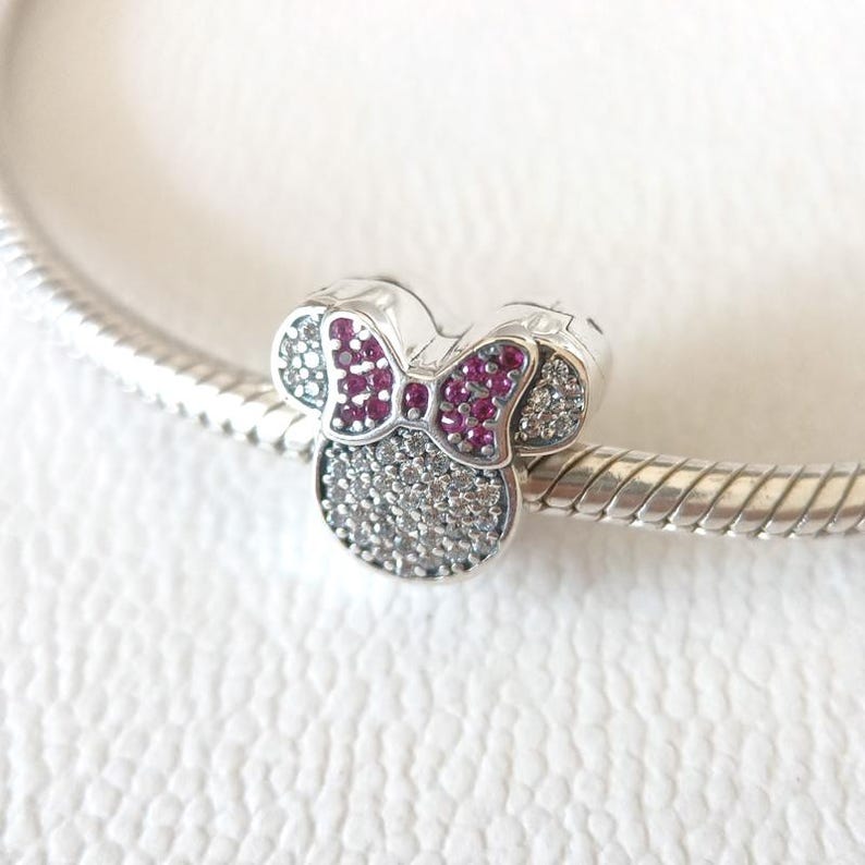 Op de afbeelding: Een zilveren bedel in de vorm van een muizenkop met een strik. De strik is rood en de kop is bedekt met kleine, heldere en rode steentjes. De bedel zit aan een zilveren armband.