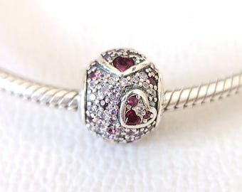 May include: A silver charm bead with a heart design. The bead is covered in small, sparkling purple and clear stones, with a larger heart-shaped design in the center. The charm is on a silver bracelet.