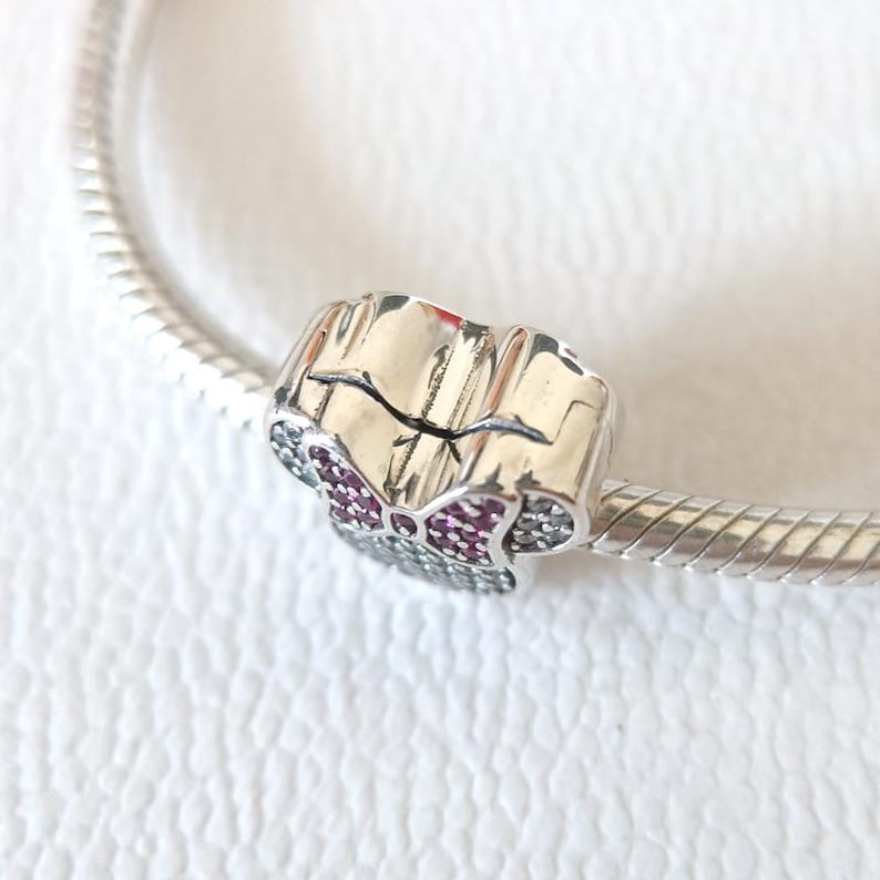 Op de afbeelding: Een zilveren bedel met een strikdesign, met een centraal gedeelte van kleine, donkerroze stenen en omringende heldere stenen. De bedel is bevestigd aan een zilveren armband, tegen een witte achtergrond.