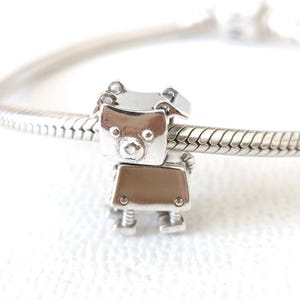 Puede incluir: Un charm plateado con forma de oso robot. El charm tiene una cabeza cuadrada con orejas pequeñas y una cara redonda. El cuerpo es rectangular con dos pequeños círculos en la parte delantera. El charm está unido a una pulsera plateada.