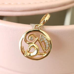 May include: A gold-coloured charm with a circular design. The charm features the letters "SA" with a decorative swirl and is embellished with small, sparkling crystals. The charm is suspended from a small loop, suggesting it is designed to be attached to a bracelet or necklace.