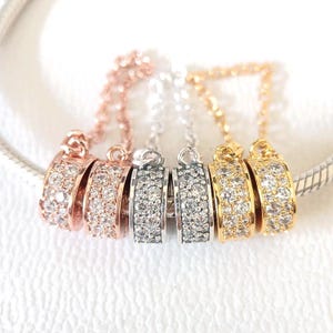 May include: Close-up of five decorative charm pendants in rose gold, silver, and gold tones. Each pendant features a curved design encrusted with small, clear gemstones. The pendants are attached to delicate chains.