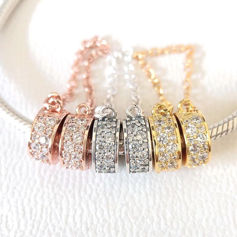 May include: Close-up of five decorative charm pendants in rose gold, silver, and gold tones. Each pendant features a curved design encrusted with small, clear gemstones. The pendants are attached to delicate chains.