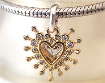 May include: A gold-toned charm with a heart-shaped center, encircled by a sunburst design with clear crystals. The heart contains the words "A DAY OF LIGHT". The charm is suspended from a silver chain.