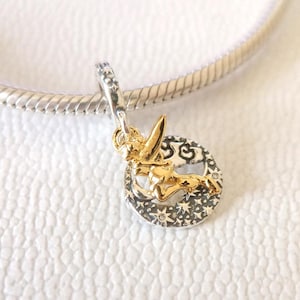 Op de afbeelding: Een zilveren bedel met een gouden feeënontwerp. De fee wordt in vlucht afgebeeld, met uitgestrekte armen en vleugels. De bedel is bevestigd aan een zilveren armband. De bedel heeft een cirkelvormig ontwerp met ster- en bloempatronen.