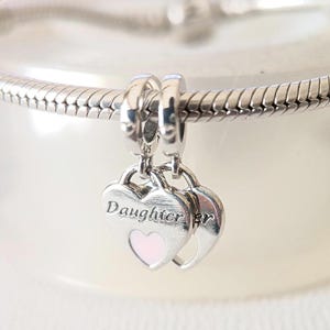 Pode incluir: Berloque de prata com dois pingentes em forma de coração. Um coração tem a palavra "Daughter" gravada e um centro em forma de coração rosa. O berloque é preso a uma pulseira de prata. Joalharia.