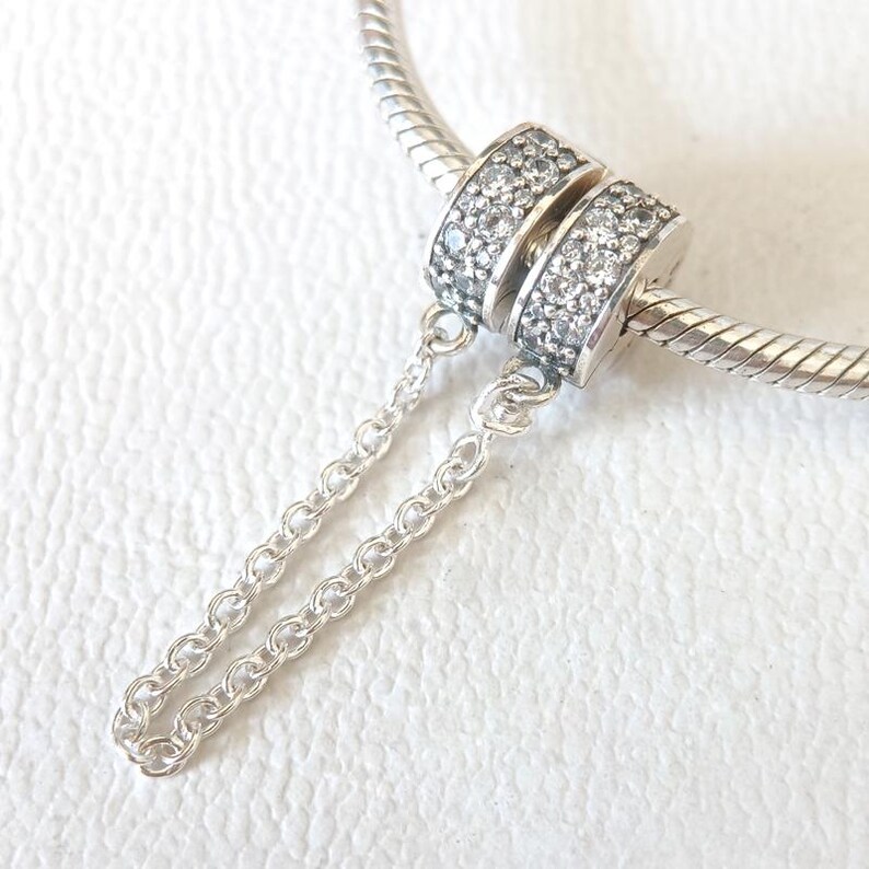 May include: A silver charm with two stacked, encrusted bands of clear stones, connected by a delicate chain. The charm is attached to a silver bracelet. The charm is a jewelry accessory.