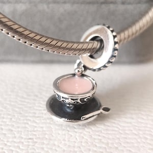 May include: A silver charm featuring a miniature teacup with a pink interior, resting on a saucer with a small spoon. The charm is attached to a silver bracelet. The teacup and saucer have decorative detailing.