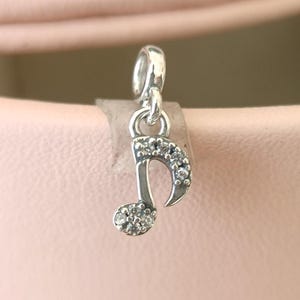 May include: A silver charm in the shape of a musical eighth note, adorned with small, clear gemstones. The charm has a loop at the top for attachment to a bracelet or necklace. The gemstones are set in a pavé style, adding sparkle to the musical note design.