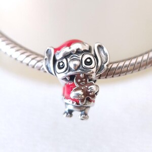 May include: A silver charm featuring a cartoon character wearing a red Santa hat and jacket, holding a gingerbread man. The charm is detailed with black eyes and a silver finish, perfect for a bracelet or necklace.
