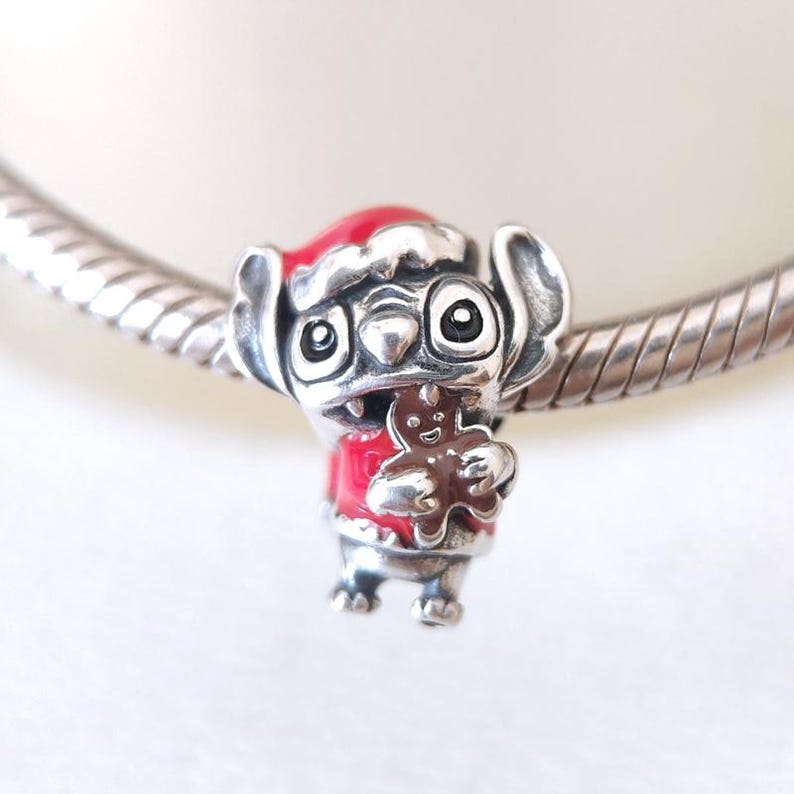 May include: A silver charm featuring a cartoon character wearing a red Santa hat and jacket, holding a gingerbread man. The charm is detailed with black eyes and a silver finish, perfect for a bracelet or necklace.