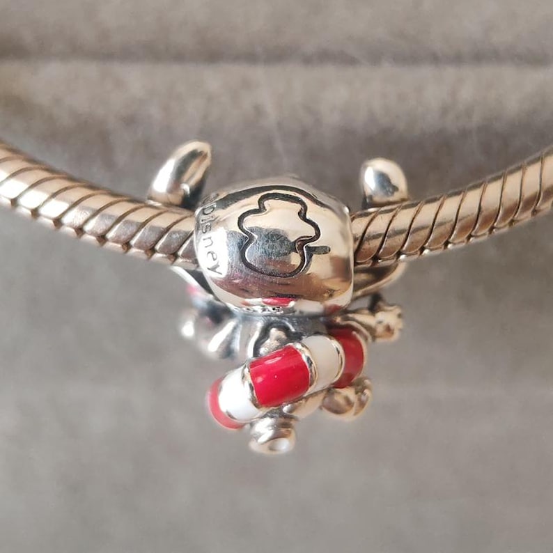 K&ouml;nnte beinhalten: Ein silberner Charm-Anh&auml;nger in Form einer Figur mit gro&szlig;en Ohren, graviert mit dem Wort Disney und einem kleeblattartigen Symbol. Die Figur h&auml;lt einen kleinen, runden Rettungsring, der mit abwechselnden roten und wei&szlig;en Emaille-Streifen verziert ist.