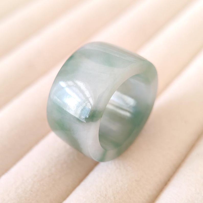K&ouml;nnte beinhalten: Ein breiter, quadratisch geformter Ring aus transluzentem Jade. Der Ring weist eine Mischung aus hellgr&uuml;nen und wei&szlig;en Farbt&ouml;nen auf, die einen marmorierten Effekt erzeugen. Die polierte Oberfl&auml;che reflektiert das Licht und hebt die nat&uuml;rlichen Variationen des Steins hervor.