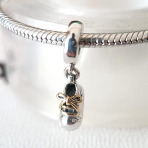 May include: A silver charm with a small baby shoe pendant. The shoe has a gold bow and is suspended from a silver bead. The charm is designed to be added to a bracelet.