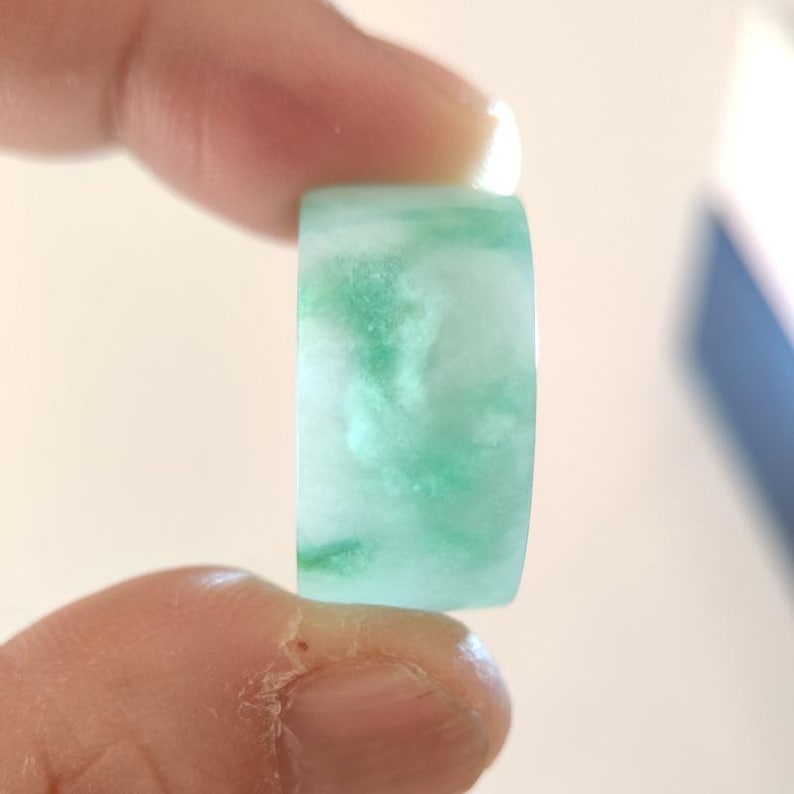 K&ouml;nnte beinhalten: Ein Jade-Ring mit marmoriertem Aussehen. Der Ring ist hellgr&uuml;n mit wei&szlig;en und dunkelgr&uuml;nen Adern. Der Ring wird zwischen zwei Fingern gehalten.