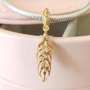 Op de afbeelding: Een goudkleurige bedel in de vorm van een veer, versierd met kleine, heldere edelstenen. De bedel hangt aan een zilverkleurige ketting. Het veermotief heeft ingewikkelde details en is een sieradenaccessoire.