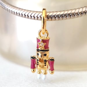 May include: A gold-colored charm featuring a miniature nutcracker soldier. The charm has a red hat with a star, pink arms, and white feet. The charm is suspended from a silver chain.