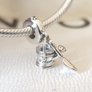 Puede incluir: Un charm de plata con temática de jardinería. Presenta una pequeña regadera con las palabras "Full Bloom" y una paleta con un diseño de mariquita. El charm está unido a una pulsera de plata.