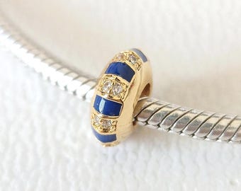 Puede incluir: Un charm dorado con detalles alternos de esmalte azul y cristales transparentes. El charm es cilíndrico y parece ser una cuenta para una pulsera. El charm está en una cadena plateada.