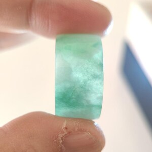 K&ouml;nnte beinhalten: Ein transluzenter Jade-Ring mit einer Mischung aus hellen und dunklen Gr&uuml;nt&ouml;nen. Der Ring wird zwischen zwei Fingern gehalten, wodurch seine glatte, polierte Oberfl&auml;che hervorgehoben wird. Der Jade hat einen wolkigen, marmorierten Effekt.