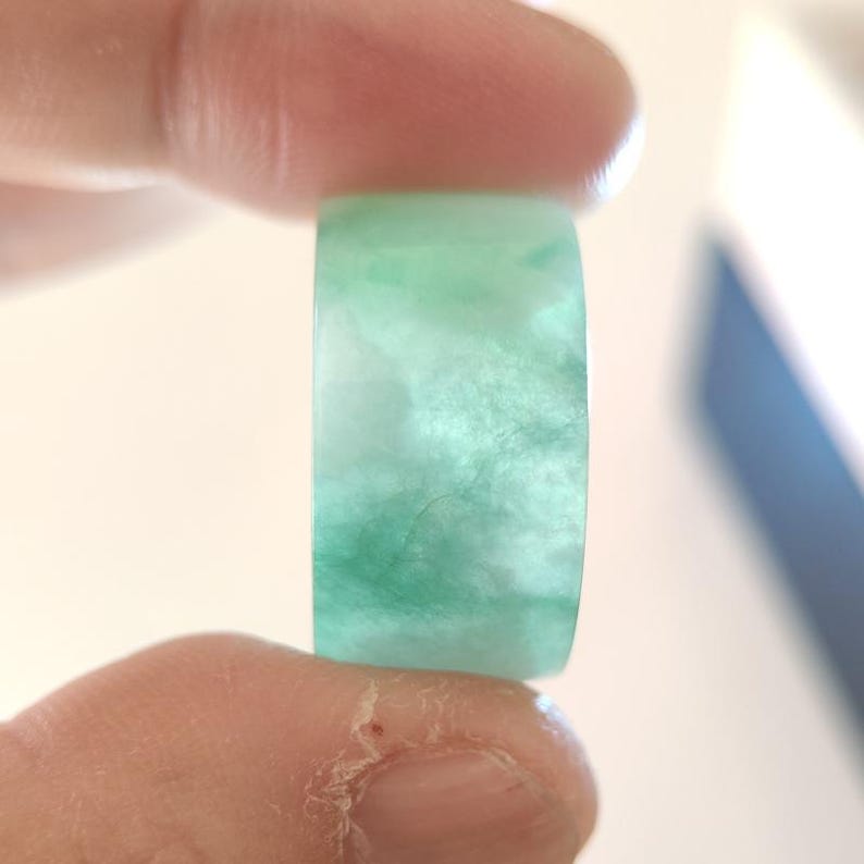 K&ouml;nnte beinhalten: Ein transluzenter Jade-Ring mit einer Mischung aus hellen und dunklen Gr&uuml;nt&ouml;nen. Der Ring wird zwischen zwei Fingern gehalten, wodurch seine glatte, polierte Oberfl&auml;che hervorgehoben wird. Der Jade hat einen wolkigen, marmorierten Effekt.