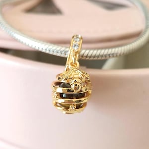 Könnte beinhalten: Ein goldfarbener Charm mit einem Bienen- und Wabenmuster. Der Charm hat oben eine Biene und darunter eine geschichtete Wabenstruktur. Der Charm ist an einem silbernen Armband befestigt. Der Charm hat kleine, klare Kristalle.