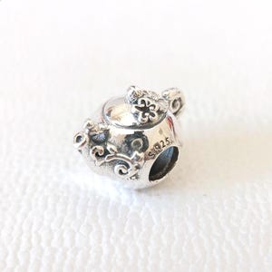 Könnte beinhalten: Ein silberner Charm in Form einer Teekanne. Der Charm hat florale und Ranken-Details, mit einer kleinen Blume auf dem Deckel. Der Charm ist mit "S925" markiert.