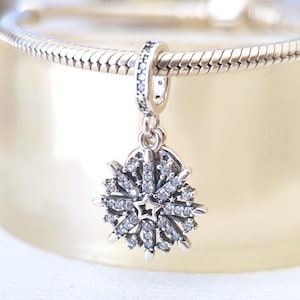 Puede incluir: Un charm de plata con forma de copo de nieve, adornado con cristales transparentes y una estrella central. El charm cuelga de una cadena de plata, ideal para una pulsera o collar. El diseño del copo es detallado y brillante.