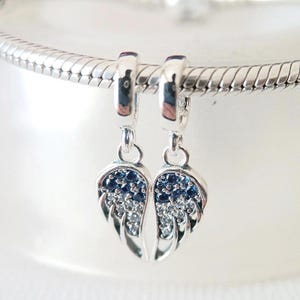 Op de afbeelding: Zilveren bedel met twee vleugelhangers. Elke vleugel is gedetailleerd met kleine, sprankelende blauwe en heldere stenen. De bedel hangt aan een zilveren ring, ontworpen voor een armband of ketting. De vleugels zijn symmetrisch.