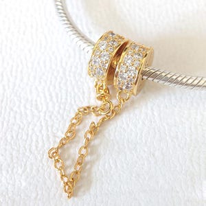 May include: Gold-toned charm with two ring-shaped elements encrusted with small, clear stones. A delicate gold chain connects the rings. The charm is displayed on a silver-colored, textured bar, against a white background.