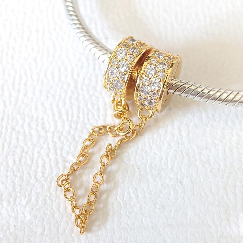 May include: Gold-toned charm with two ring-shaped elements encrusted with small, clear stones. A delicate gold chain connects the rings. The charm is displayed on a silver-colored, textured bar, against a white background.
