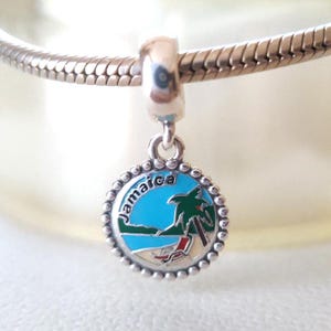Può includere: Charm in argento con un ciondolo rotondo raffigurante una scena di spiaggia. Il ciondolo ha un cielo blu, una palma verde e una sedia a sdraio rossa e bianca. La parola "Jamaica" è scritta in bianco.