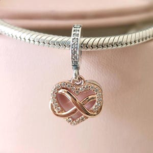 Könnte beinhalten: Ein roségoldener Charm-Anhänger mit herzförmigem Design, mit einem Unendlichkeitssymbol und mit klaren Kristallen besetzt. Der Charm hängt an einem silbernen Armband. Das Herz hat eine rosa Mitte.