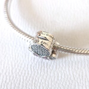 Op de afbeelding: Een zilveren bedel met het woord "Disney" gegraveerd. De bedel is versierd met kleine, glinsterende steentjes en is bevestigd aan een zilveren armband. De bedel is een sieraad.