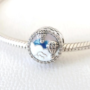 Puede incluir: Un charm de plata con un diseño de dos globos aerostáticos flotando sobre nubes blancas y cielo azul. El charm es redondo, con un borde de pequeñas piedras transparentes. El fondo es blanco y el charm está en una pulsera de plata.