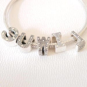 May include: A silver charm bracelet with the word "SUWITI" spelled out in individual letter charms. Each charm is detailed with small heart-shaped cutouts. The bracelet is on a white background.