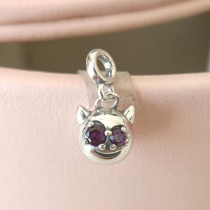Może przedstawiać: Srebrny charms z motywem kociej twarzy. Charms posiada dwa okrągłe, ciemnofioletowe kamienie szlachetne jako oczy i uśmiechnięte usta. Charms ma małe, spiczaste uszy i pętlę do mocowania.