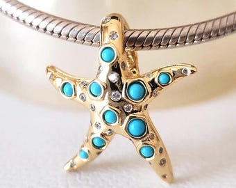 Puede incluir: Un colgante de estrella de mar dorado con detalles en azul turquesa y pequeñas gemas transparentes. El colgante está sujeto a una cadena plateada. El diseño de estrella de mar es un tema popular para la joyería.