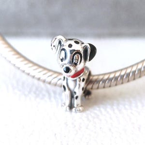 Puede incluir: Un charm plateado con forma de perro dálmata. El charm tiene manchas negras, orejas negras y un collar rojo. El perro está sentado con las patas delanteras extendidas. El charm está en una pulsera plateada.