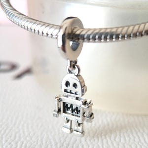 May include: A silver robot charm suspended from a silver bracelet. The robot features a detailed design with a square head, rectangular body, and articulated limbs. The charm is made of a shiny metal.
