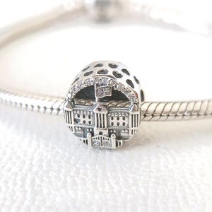 May include: A silver charm with a detailed architectural design, featuring a building with a flag and sparkling accents. The charm is round with a patterned top and is displayed on a silver bracelet.