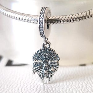 Könnte beinhalten: Ein silberner Charm-Anhänger mit dem Design des Raumschiffs Millennium Falcon. Der Anhänger hängt an einem silbernen Ring mit kleinen, klaren Steinen. Der Charm ist detailliert und an einer silbernen Kette befestigt.