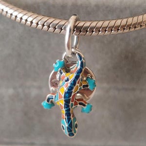 Puede incluir: Un dije de metal plateado con forma de lagartija, que presenta una incrustación de esmalte estilo mosaico en tonos azul brillante, amarillo y naranja. El dije de lagartija cuelga de una argolla plateada sujeta a una pulsera de cadena tipo serpiente.